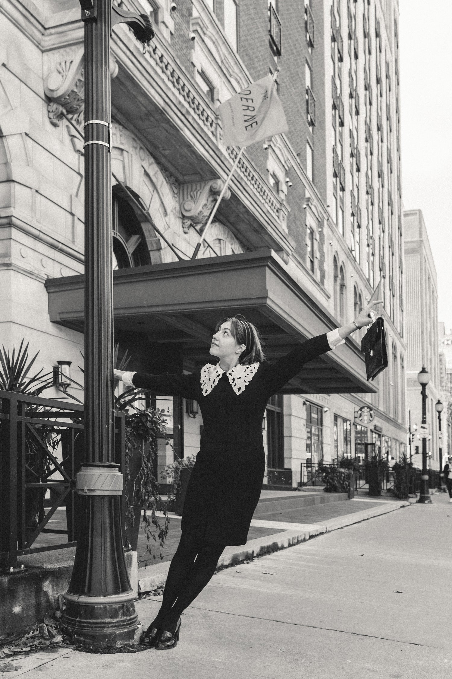 Vintage girl hangs on a lamp post in a black dress with large cream lace collar. Old architecture surrounds her.